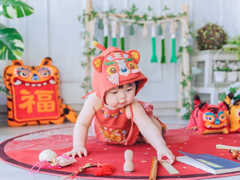 寶貝坐在紅色抓周墊上,從擺好的道具裡伸手抓取